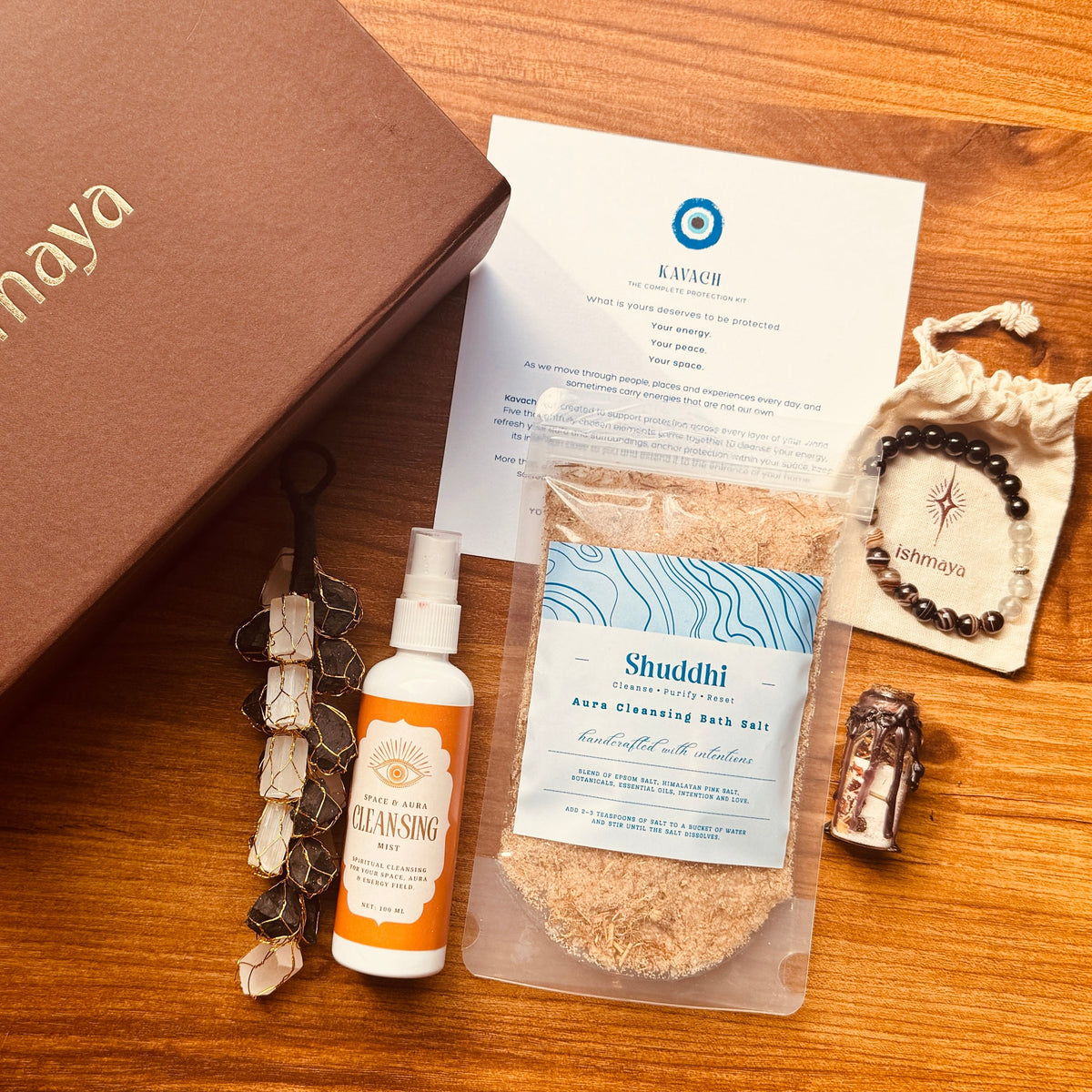 A curated wellness gift set arranged on a warm wooden tabletop, featuring a brown branded box, a packet of bath salts, a small spray bottle, a card, a beaded bracelet, and a small jar.