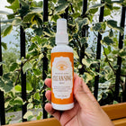 A hand holding a white spray bottle of Space & Aura Cleansing Mist against a background of green and cream variegated houseplant leaves and a black metal fence.